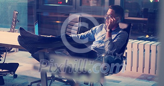 Full length of a relaxed casual young businessman sitting with legs on desk at night office Full length of a relaxed casual young businessman sitting with legs on desk at night office