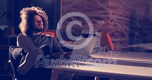 Full length of a relaxed casual young businessman sitting with legs on desk at night office Full length of a relaxed casual young businessman sitting with legs on desk at night office