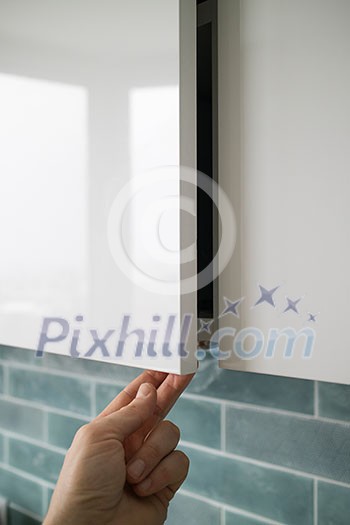 The closeup man hand opening or closing kitchen cabinet door on a background of blue kitchen interior. Modern kitchen furniture without handles with an additional small lower for opening the door The closeup man hand opening or closing kitchen cabinet door on a background of blue kitchen interior. Modern kitchen furniture without handles with an additional small lower for opening the door