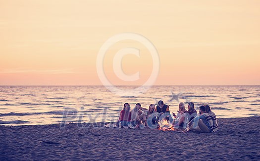 Group of young friends sitting by the fire at autumn beach, grilling sausages and drinking beer, talking and having fun