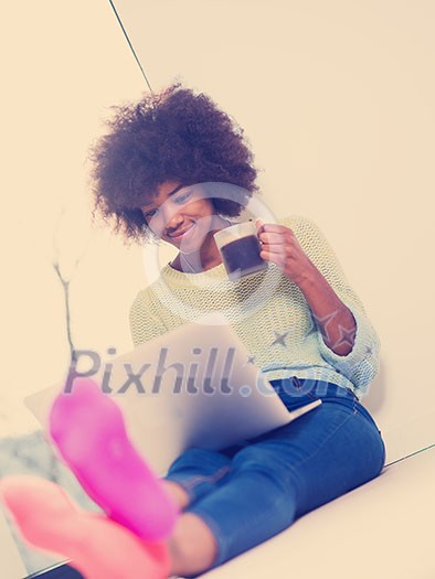 Young african american woman smiling sitting on the floor near bright window while looking at open laptop computer and holding mug at home Young african american woman smiling sitting on the floor near bright window while looking at open laptop computer and holding mug at home