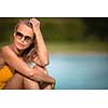 Young Beautiful Suntanned Woman wearing sunglasses relaxing next to a Swimming Pool  on a lovely Summer Day