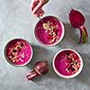 The girl's hand puts oatmeal in a plate with freshly prepared beetroot smoothies with raspberries and currants on a gray concrete background. Top view. Concept of diet and vegetarian food