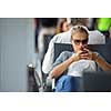 Casual blond young woman using her cell phone while waiting to board a plane at departure gates at international airport.