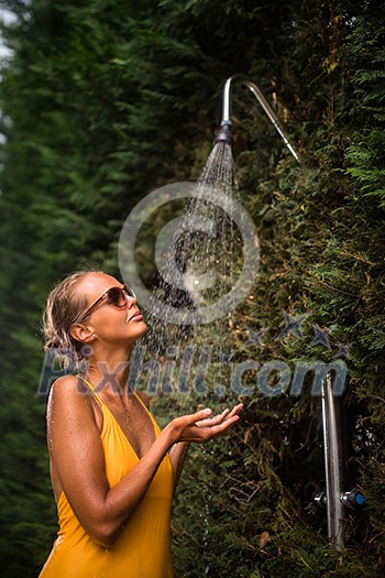 Beautiful young woman taking a shower outdoors. Attractive young woman in yellow one-piece enjoys shower on hot sunny day after a swim. Heat and refreshment concept.