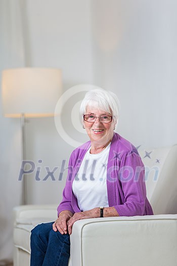 Portrait of a senior woman at home - Looking happy, looking at the camera, smiling while sitting on the sofa in her living room Portrait of a senior woman at home - Looking happy, looking at the camera, smiling while sitting on the sofa in her living room