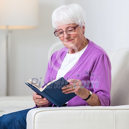 Portrait of a senior woman at home - Looking happy, looking at the camera, smiling while sitting on the sofa in her living room and reading a good book Portrait of a senior woman at home - Looking happy, looking at the camera, smiling while sitting on the sofa in her living room and reading a good book