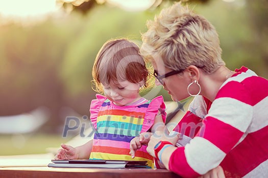 Happy mother and her little daughter enjoying free time using tablet computer while relaxing on holiday home garden during sunny day Happy mother and her little daughter enjoying free time using tablet computer while relaxing on holiday home garden during sunny day