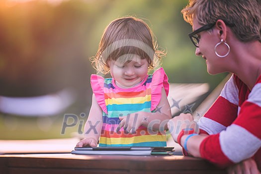 Happy mother and her little daughter enjoying free time using tablet computer while relaxing on holiday home garden during sunny day Happy mother and her little daughter enjoying free time using tablet computer while relaxing on holiday home garden during sunny day