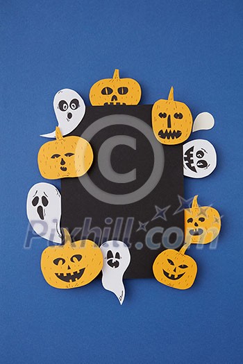 Greeting decorative card handcraft with paper horrible smiling spirits and ghosts and laughing pumpkin on a blue paper background, place under text. Top view. Greeting decorative card handcraft with paper horrible smiling spirits and ghosts and laughing pumpkin on a blue paper background, place under text. Top view.