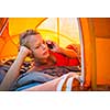 Pretty, young woman camping outdoors, lying in the tent in the morning, using her smart phone
