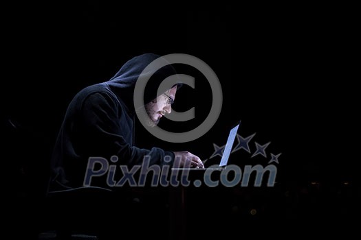 Young talented hacker using laptop computer while working in dark office with big city lights in the background at night Young talented hacker using laptop computer while working in dark office with big city lights in the background at night