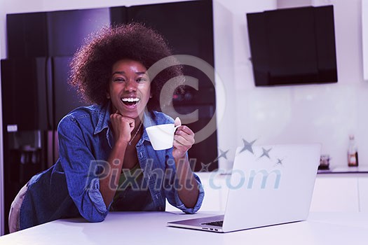 Young smiling black woman using computer and drinking coffee in modern kitchen interior Young smiling black woman using computer and drinking coffee in modern kitchen interior