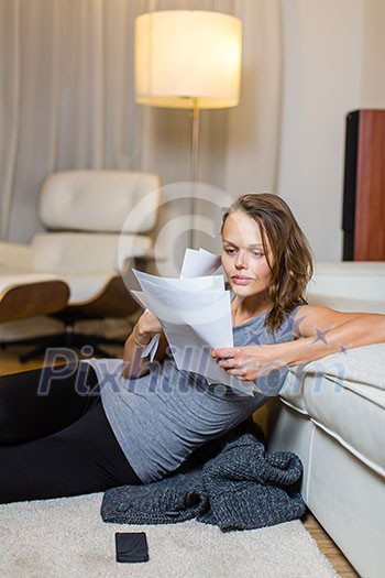 Pretty, young woman going through some paperwork in her lovely living room at night, looking rather tired Pretty, young woman going through some paperwork in her lovely living room at night, looking rather tired