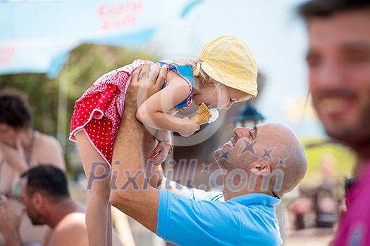 cute little girl having fun with her young father while eating ice cream by the sea during summer vacation holidays, celebration, children and people concept