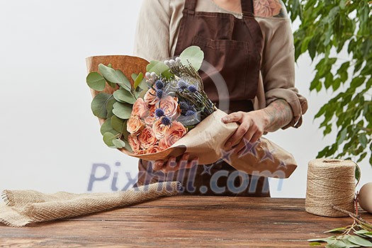 Gardener woman with bouquet for a holiday from living coral roses and branches of green leaf on a wooden table on a light background, place for text. Mother's Day holiday. Gardener woman with bouquet for a holiday from living coral roses and branches of green leaf on a wooden table on a light background, place for text. Mother's Day holiday.