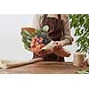Gardener woman with bouquet for a holiday from living coral roses and branches of green leaf on a wooden table on a light background, place for text. Mother's Day holiday.