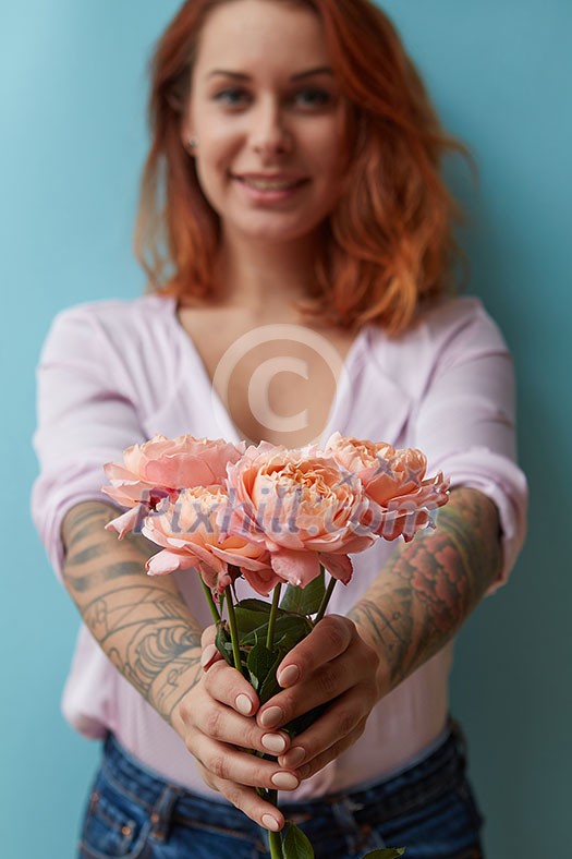 A happy red-haired woman with a tattoo holds in her hands delicate pink roses on a blue background with copy space for text. Holiday gift concept A happy red-haired woman with a tattoo holds in her hands delicate pink roses on a blue background with copy space for text. Holiday gift concept