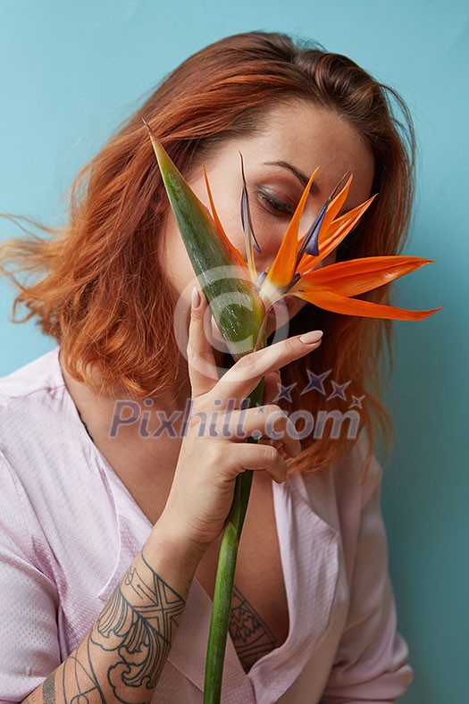 Woman with a tattoo holding an exotic orange flower Strelitz in hands around a blue background with space for text. Valentine's day concept Woman with a tattoo holding an exotic orange flower Strelitz in hands around a blue background with space for text. Valentine's day concept