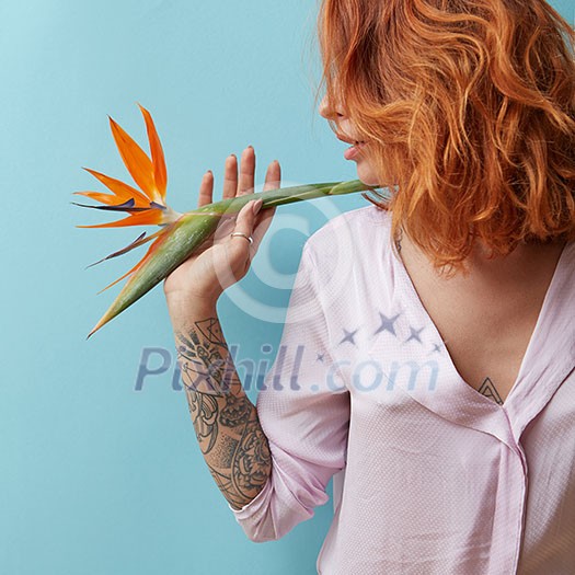 Sexy, red-haired woman with a tattoo holding an exotic strelitzia flower on a blue background with copy space. Valentine's day card Sexy, red-haired woman with a tattoo holding an exotic strelitzia flower on a blue background with copy space. Valentine's day card