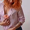 Pretty woman with ginger hair, tattoo on her bust and hands holds fresh rose living coral color on a light background. Copy space. Postcard for Woman's or Mother's Day.