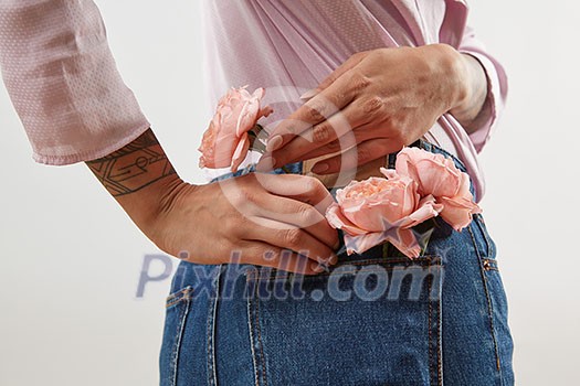 Young girl wearing of jeans pants and pink shirt from back put in natural flowers roses in a pocket on a gray background, place for text. Concept of Woman's or Mother's Day. Young girl wearing of jeans pants and pink shirt from back put in natural flowers roses in a pocket on a gray background, place for text. Concept of Woman's or Mother's Day.