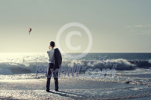 Young relaxed businessman at seaside talking mobile and looking far away at sea. Mixed media Young relaxed businessman at seaside talking mobile and looking far away at sea. Mixed media