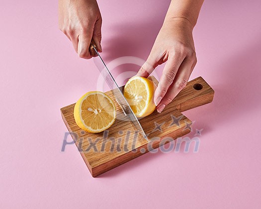 On a wooden board the hands of a woman cut a ripe lemon for making a vitamin drink around a pink background with space for text. On a wooden board the hands of a woman cut a ripe lemon for making a vitamin drink around a pink background with space for text.