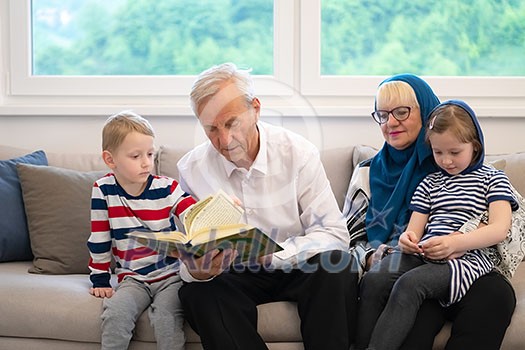 modern muslim family grandparents with grandchildren reading Quran and praying together on the sofa before iftar dinner during a ramadan feast at home modern muslim family grandparents with grandchildren reading Quran and praying together on the sofa before iftar dinner during a ramadan feast at home