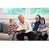 modern muslim family grandparents with grandchildren reading Quran and praying together on the sofa before iftar dinner during a ramadan feast at home