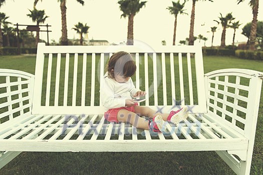 beautiful little girl playing with mobile phone while sitting on the bench in the park beautiful little girl playing with mobile phone while sitting on the bench in the park