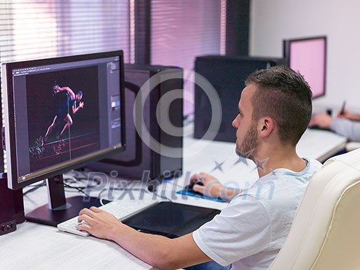 Young Graphic Designer Working at Workplace. Guy Sitting at Table Retouching Photo Using Digital Tablet with Pen and desktop computer Indoors. Freelancing and Distance Job Concept Young Graphic Designer Working at Workplace. Guy Sitting at Table Retouching Photo Using Digital Tablet with Pen and desktop computer Indoors. Freelancing and Distance Job Concept