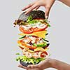 Homemade delicious sandwich from flying ingredients shrimp, fresh organic vegetables and black bun in a woman's hands on a light grey background with copy space.