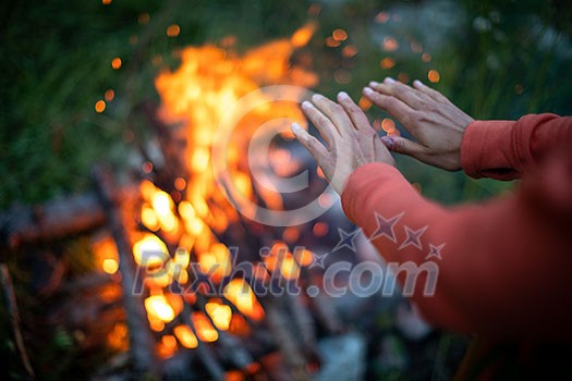 Young woman making fire while camping outdoors, in an alpine wilderness - warming up her hands near the fire Young woman making fire while camping outdoors, in an alpine wilderness - warming up her hands near the fire