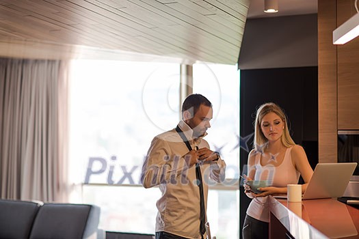 A young couple is preparing for the job and using a laptop. The man drinks coffee while the woman eats breakfast at luxury home together, looking at screen, smiling. A young couple is preparing for the job and using a laptop. The man drinks coffee while the woman eats breakfast at luxury home together, looking at screen, smiling.