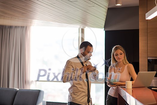 A young couple is preparing for the job and using a laptop. The man drinks coffee while the woman eats breakfast at luxury home together, looking at screen, smiling.