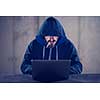 Young talented hacker smoking cigarette while working on laptop computer in dark office with concrete wall in the background