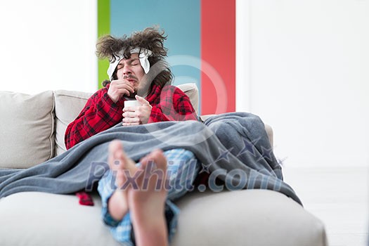 young Man in bathrobe with flu and fever wrapped holding cup of healing tea while sitting on sofa at home young Man in bathrobe with flu and fever wrapped holding cup of healing tea while sitting on sofa at home