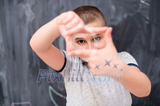 happy cute little boy having fun making hand frame gesture while standing in front of black chalkboard happy cute little boy having fun making hand frame gesture while standing in front of black chalkboard