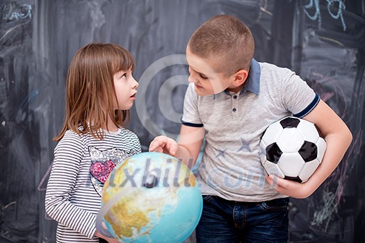 happy childrens boy with soccer ball and little girl learning about the world using globe of earth while standing in front of black chalkboard happy childrens boy with soccer ball and little girl learning about the world using globe of earth while standing in front of black chalkboard