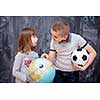 happy childrens boy with soccer ball and little girl learning about the world using globe of earth while standing in front of black chalkboard