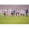 group of young professional american football players with coach discussing strategy together during training match on the stadium field