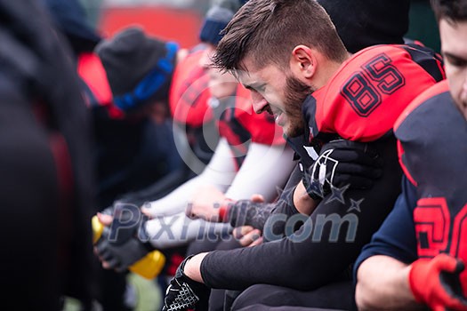 A tired american football player holding the injured shoulder while resting with teammates after hard training at field A tired american football player holding the injured shoulder while resting with teammates after hard training at field