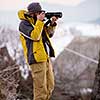 Young cheerful male photographer taking photographs of winter forest with digital camera at mountains  Young man shooting photos outdoors