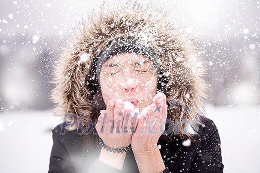 Happy young woman wearing winter clothes while blowing snow on snowy day with snowflakes around her in beautiful winter forest Happy young woman wearing winter clothes while blowing snow on snowy day with snowflakes around her in beautiful winter forest