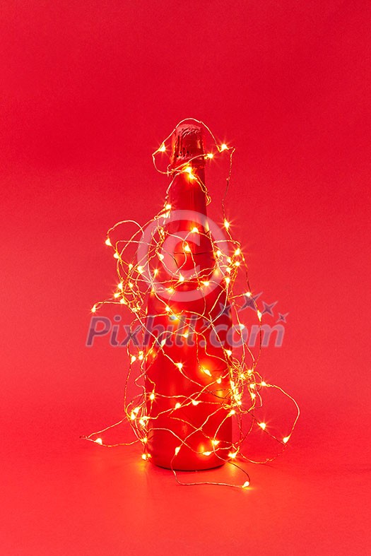 Shined Christmas lights string on a creative painted wine bottle on a red background with copy space. New Year congratulation card. Shined Christmas lights string on a creative painted wine bottle on a red background with copy space. New Year congratulation card.