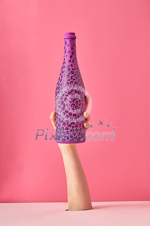 Woman's hand holds painted bottle with black spots on a pink background, copy space. Woman's hand holds painted bottle with black spots on a pink background, copy space.