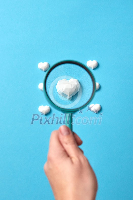 Woman's hand holds big magnifying glass above focused plaster figure of heart among others of pattern on a pastel blue background, copy space. Valentine's day greeting card. Woman's hand holds big magnifying glass above focused plaster figure of heart among others of pattern on a pastel blue background, copy space. Valentine's day greeting card.