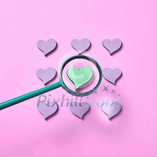 Big loupe is increesing size of green heart around other lavender colored hearts on a pastel pink background, copy space. Valentine's day greeting card. Big loupe is increesing size of green heart around other lavender colored hearts on a pastel pink background, copy space. Valentine's day greeting card.