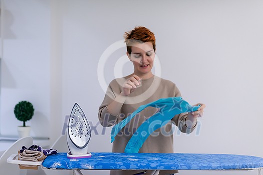 beautiful young red haired housewife in good mood ironing clothes on ironing board in front of a white wall at home beautiful young red haired housewife in good mood ironing clothes on ironing board in front of a white wall at home